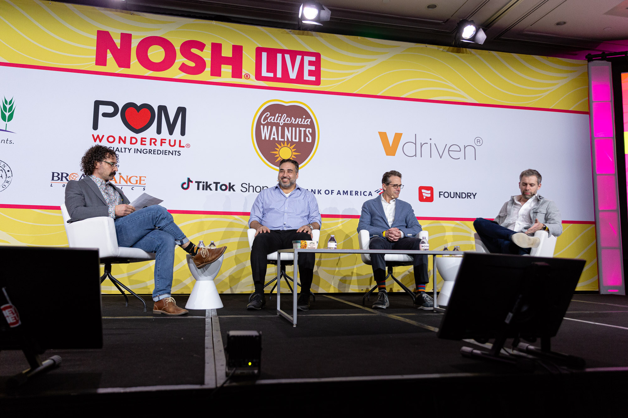 Wide view of four men seated in white chairs on a stage with a yellow Nosh Live backdrop and sponsor logos.