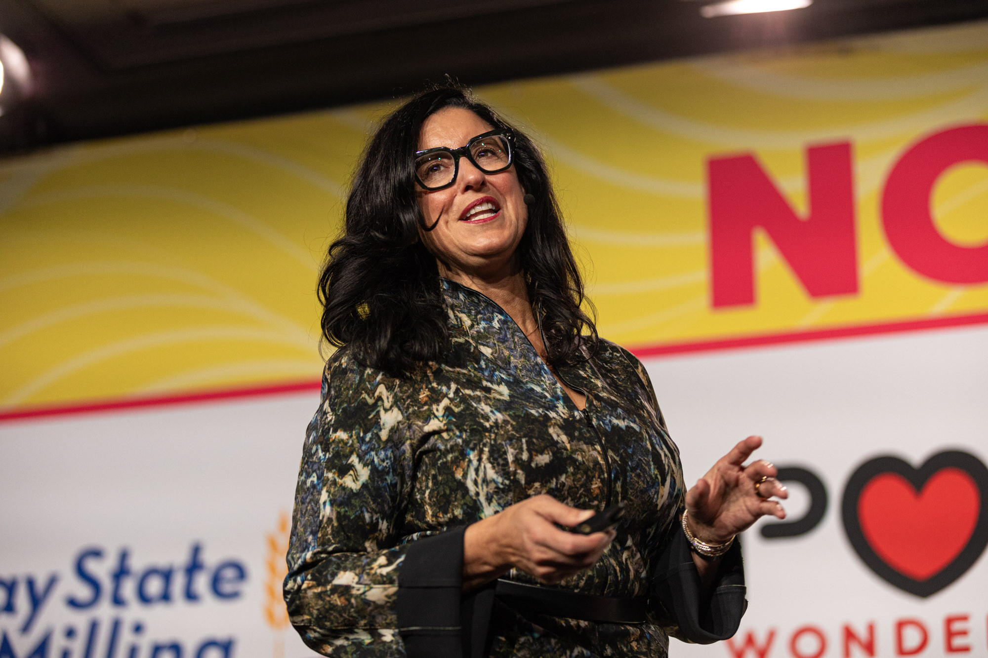 A woman in a patterned dress and glasses presenting on a stage in front of a screen.