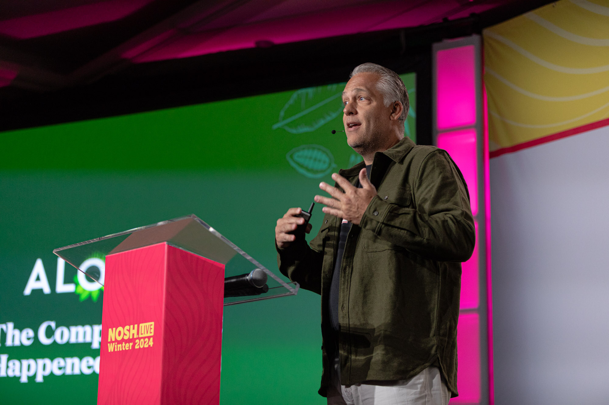 A man with slicked-back silver hair in an olive overshirt speaking at a podium, with a green screen behind him.