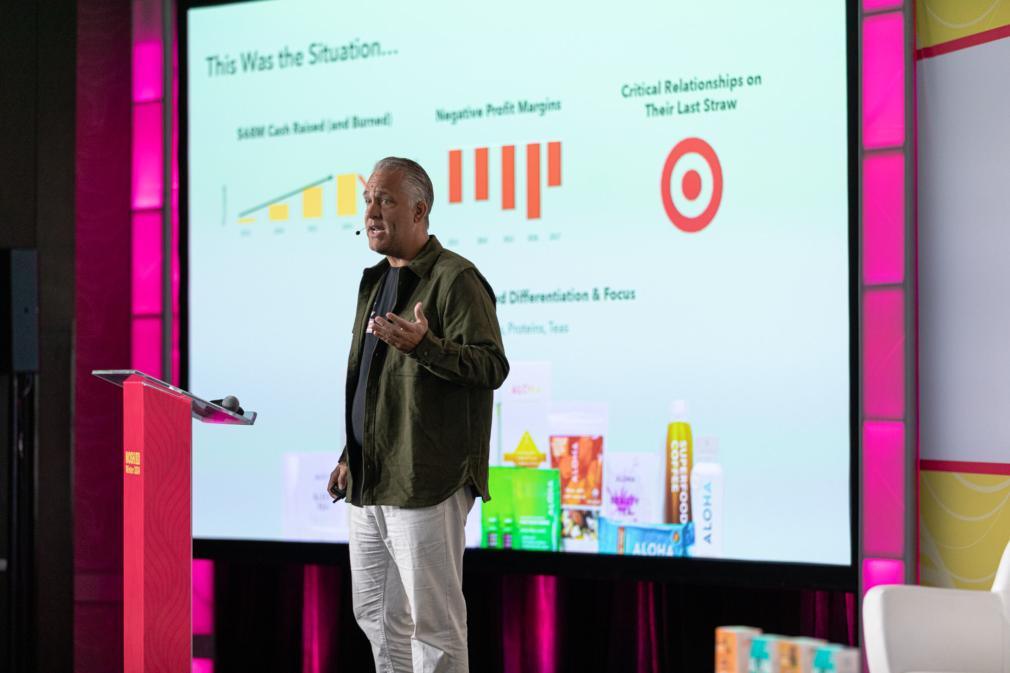 A man in an olive jacket and white pants standing on stage beside a large slide titled This Was the Situation with product packages.