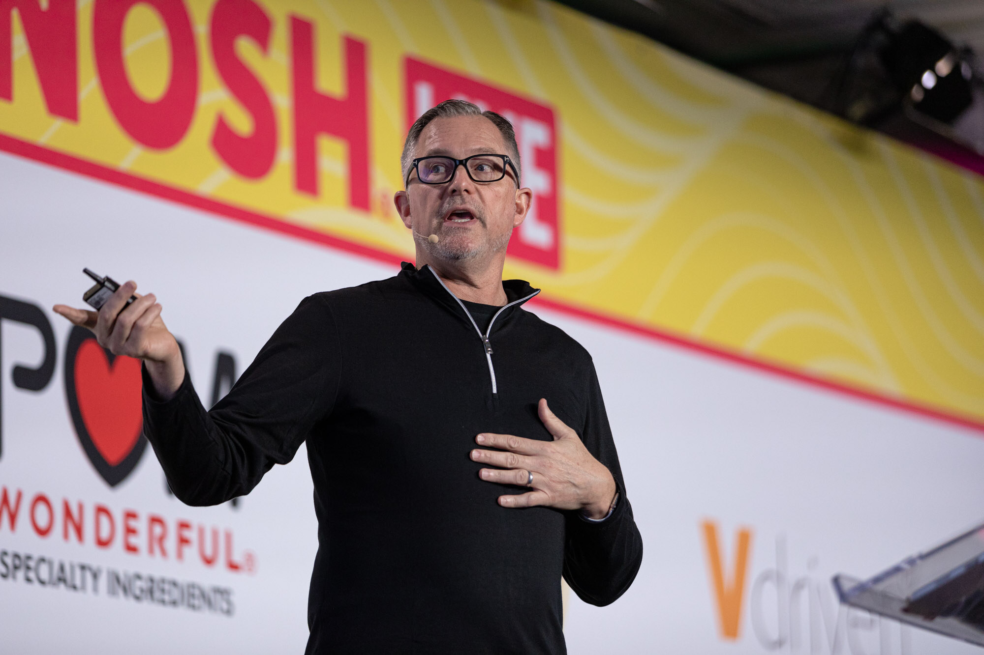 A man in a black quarter-zip top and dark-rimmed glasses speaking with a hand across his chest.