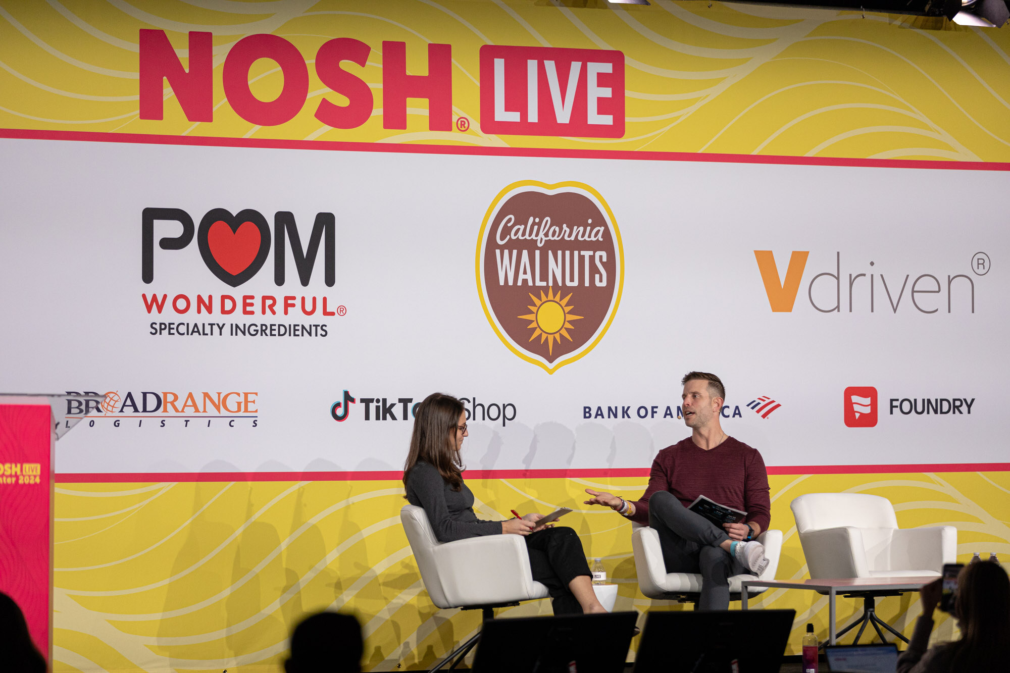 Wide view of a man in a maroon sweater and a woman in gray seated in chairs in front of the Nosh Live backdrop.