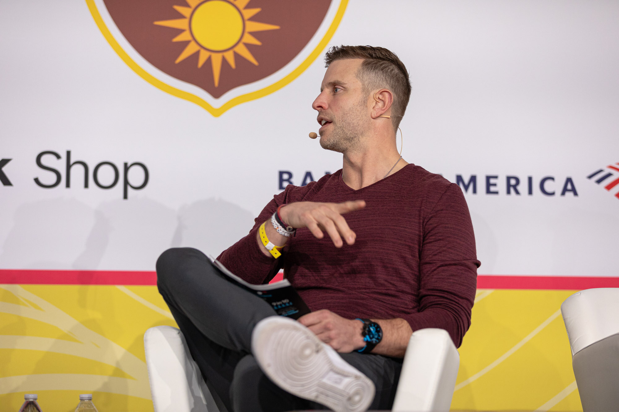 A man in a maroon V-neck sweater and white sneakers gesturing with one hand, seated in front of sponsor logos.