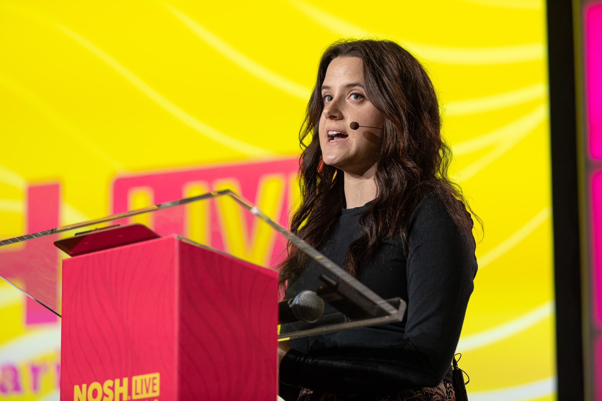 A woman with long dark hair in a dark top speaking at a red Nosh Live podium.