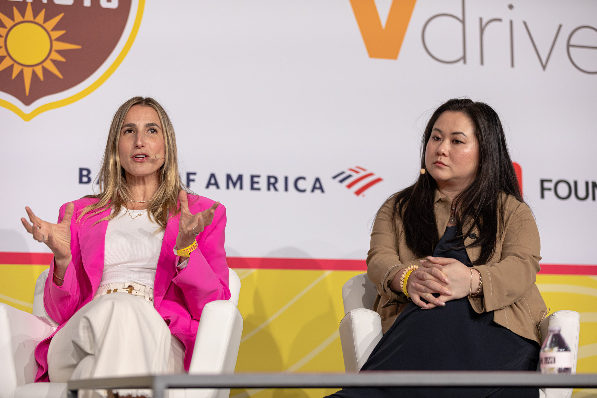 A woman in a hot-pink blazer and white outfit seated beside a woman in a tan jacket, both listening.