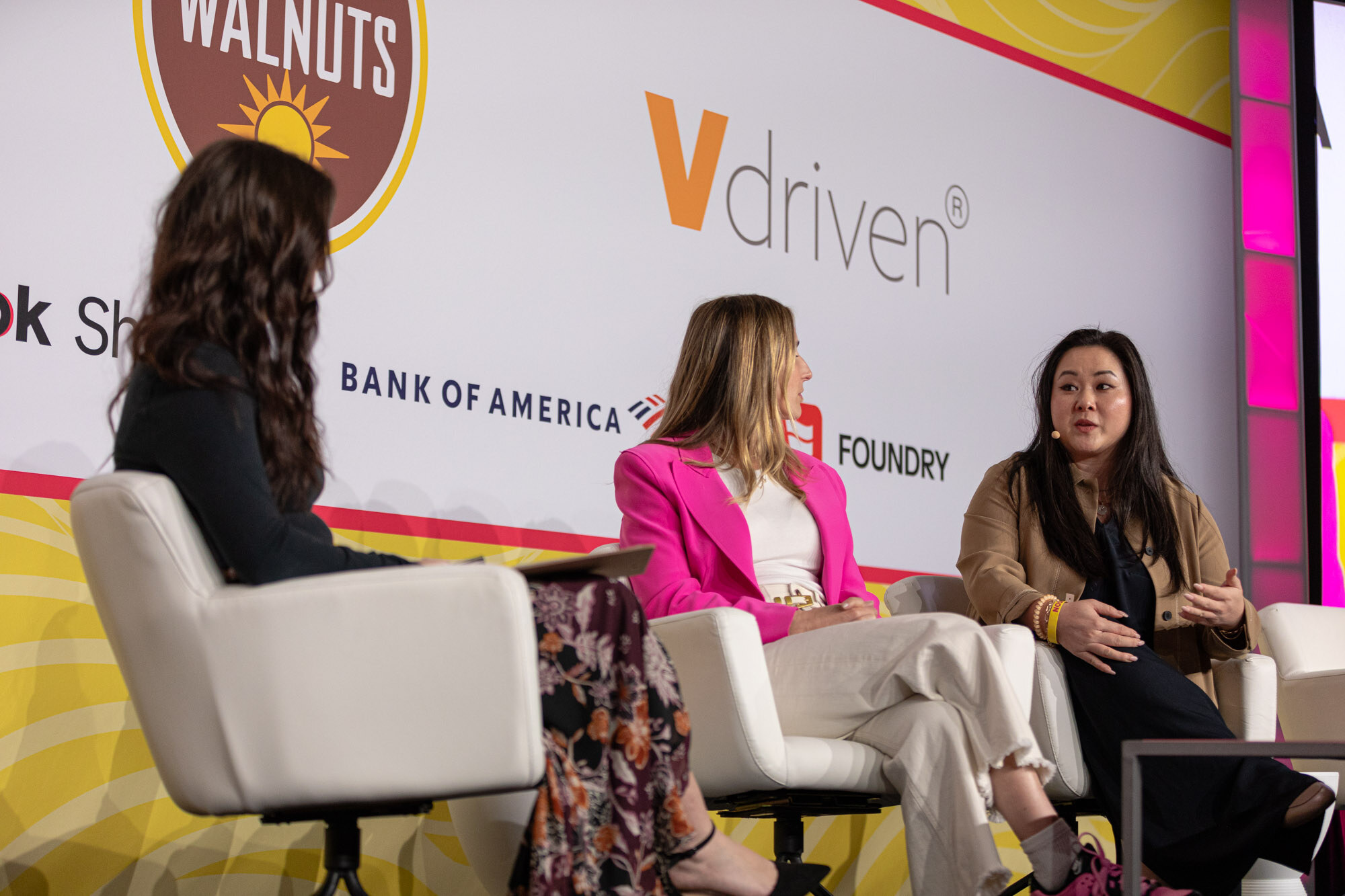 Three women seated in white chairs, one in a floral skirt, one in a hot-pink blazer and one in a tan jacket.