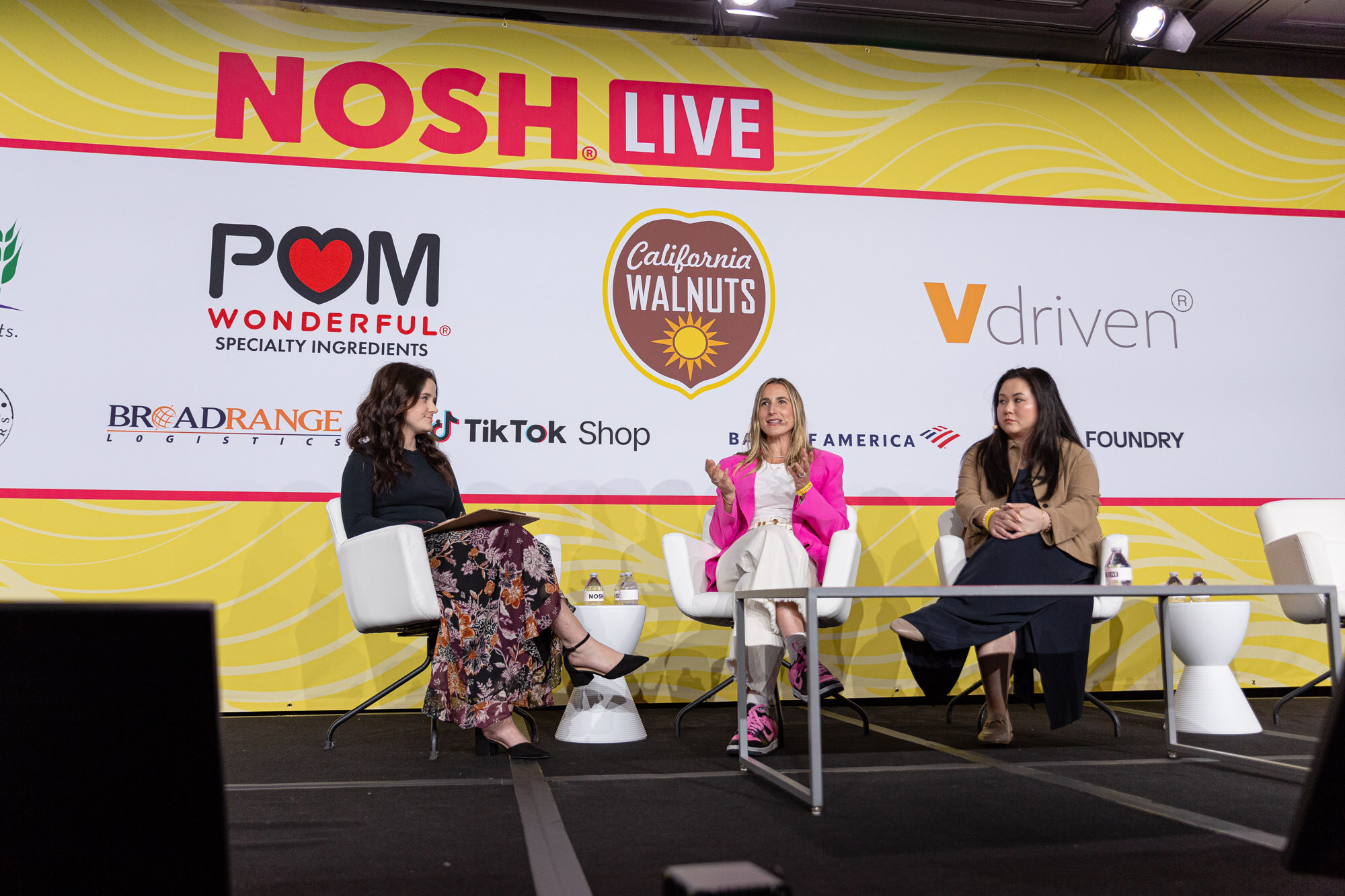 Wide view of three women seated on a stage, one in a pink blazer laughing, in front of sponsor logos.