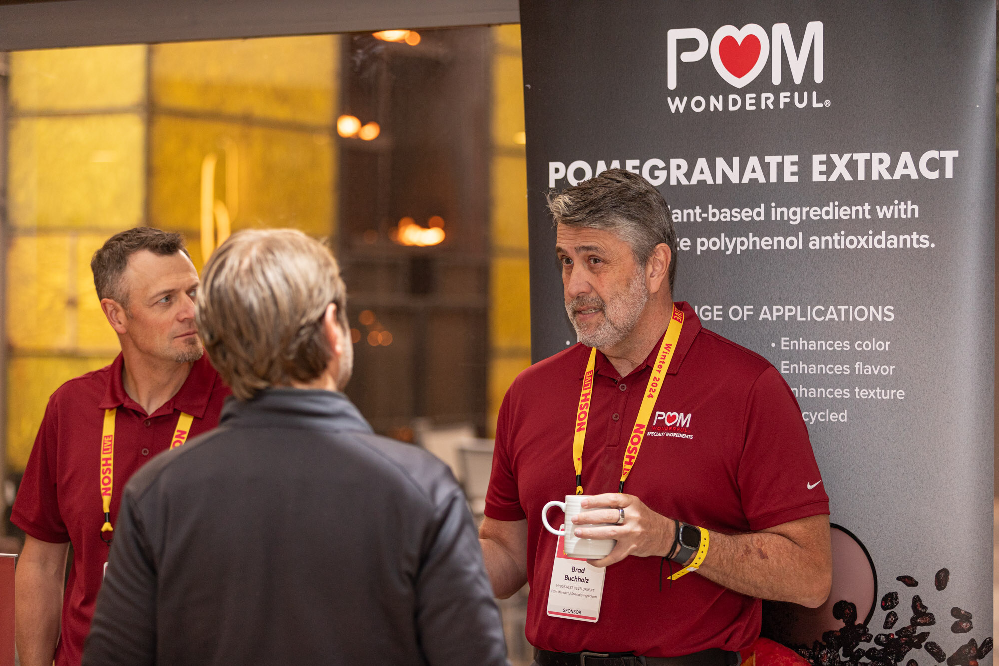Two men in red polo shirts and an attendee in front of a banner reading POM Wonderful Pomegranate Extract.