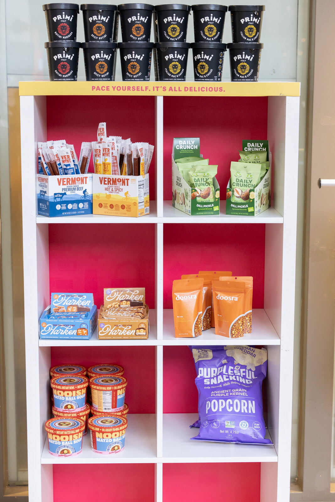 A white shelf with pink back panels stocked with snack bags, cups and jerky packs.