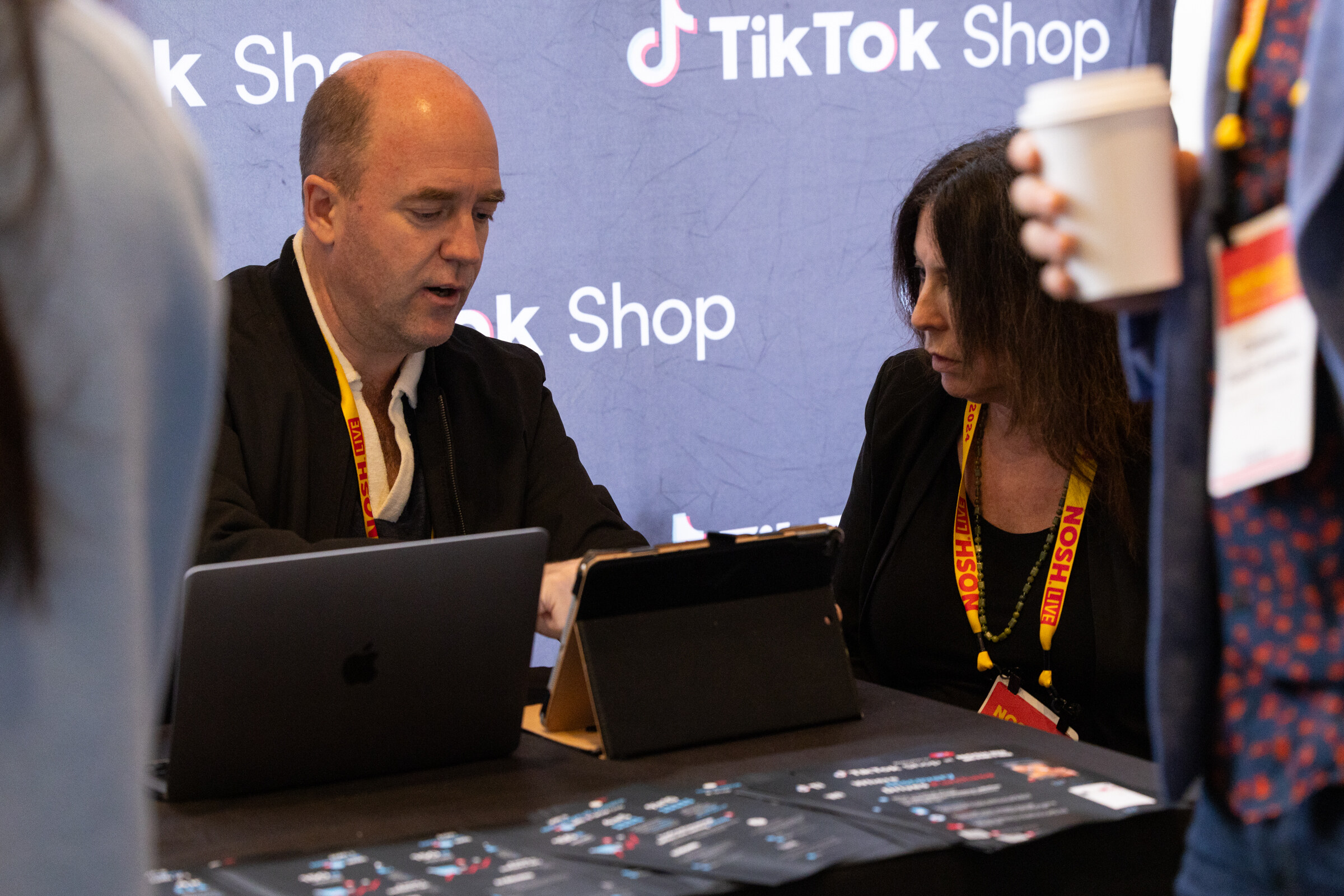 A man and a woman with laptops at a table in front of a TikTok Shop backdrop.