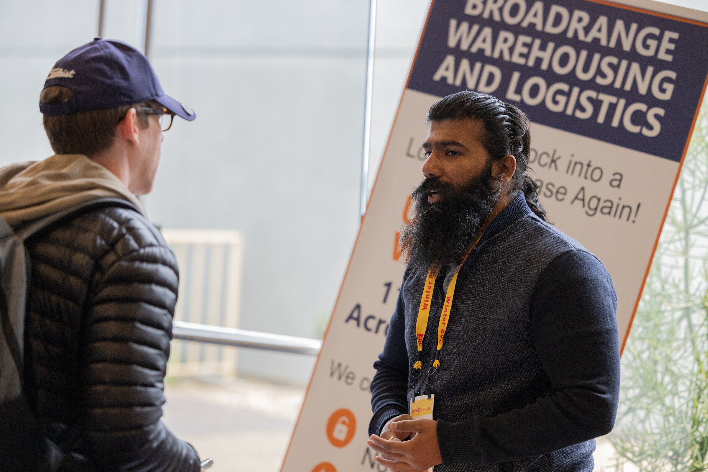 A bearded man in a navy vest with a lanyard in front of a banner reading Broadrange Warehousing and Logistics.