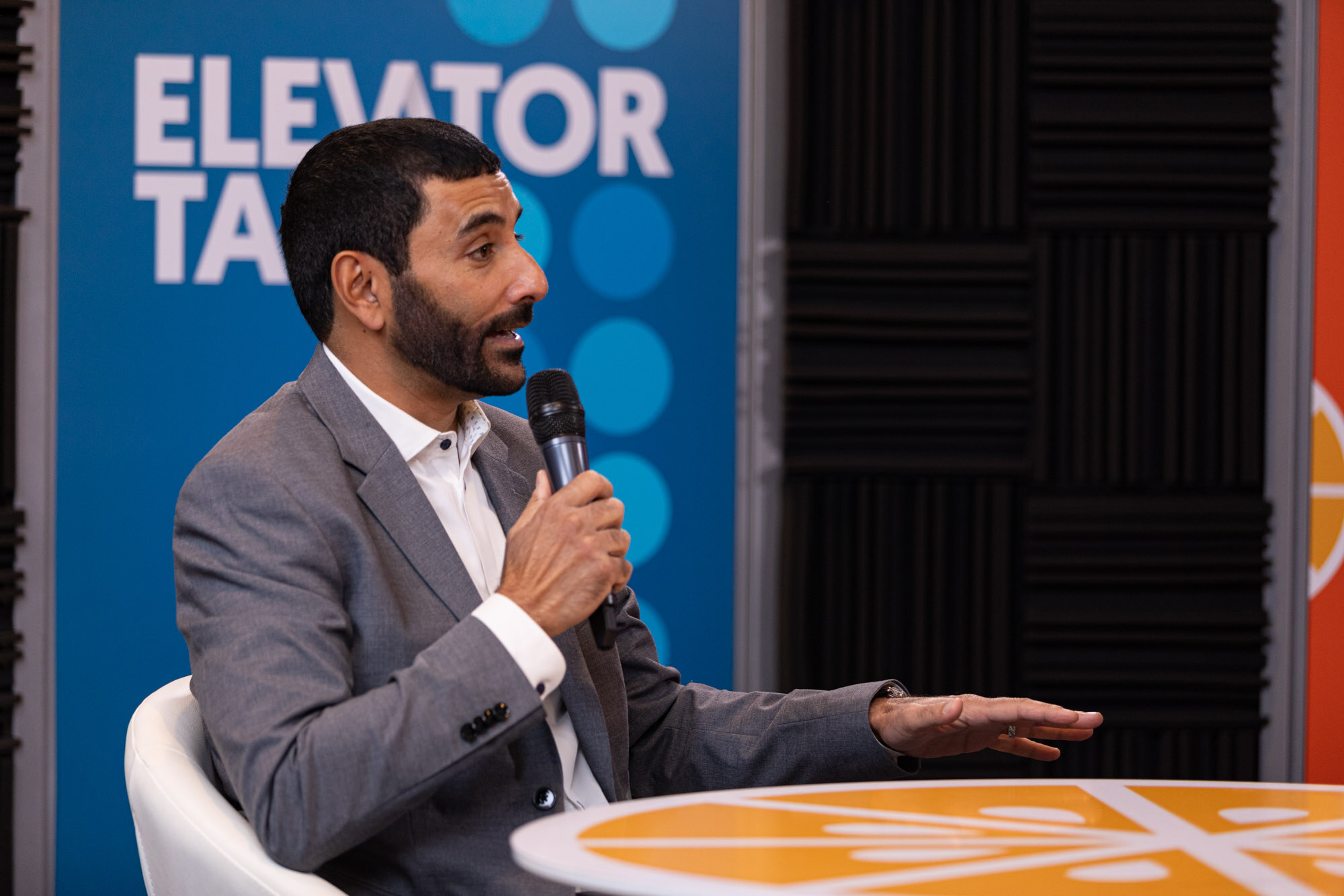 A man in a gray blazer holding a microphone in front of a blue banner reading Elevator Talk.
