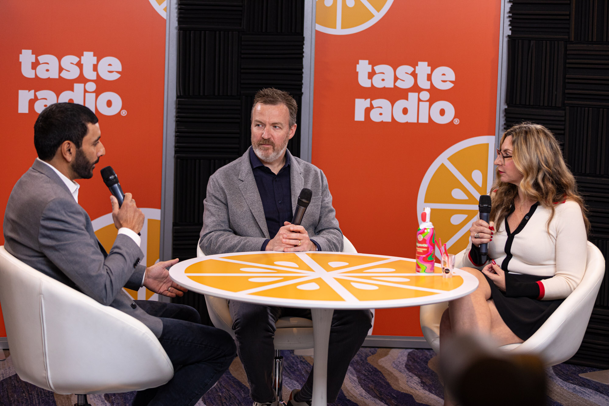 A man in a gray blazer, a man in a gray jacket and a blonde woman at an orange-slice table in front of a Taste Radio banner.