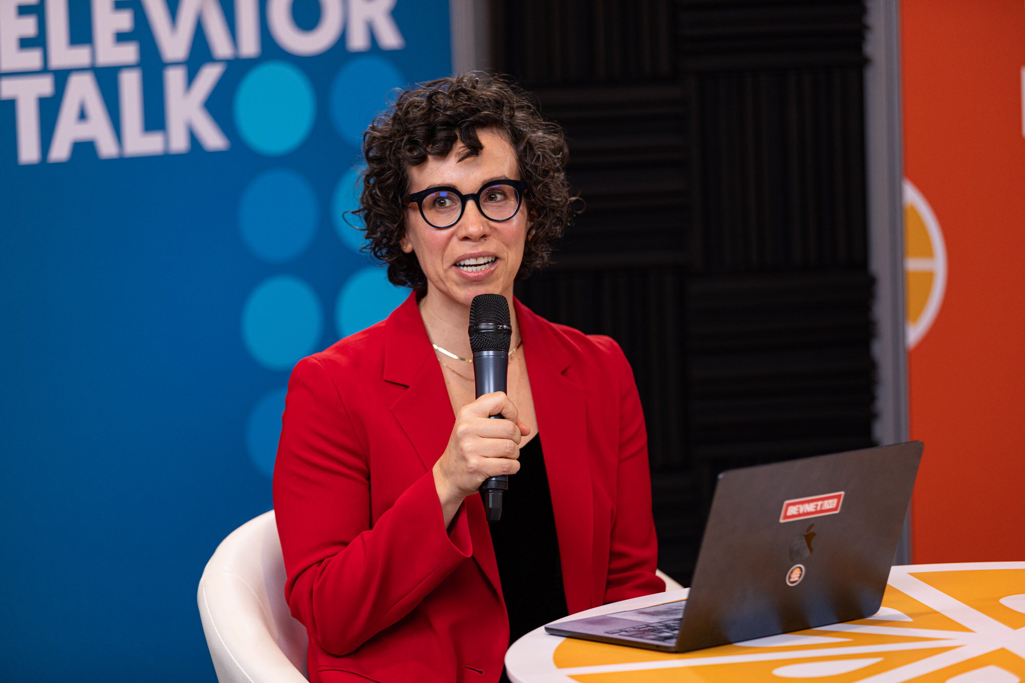 A woman with curly dark hair, round glasses and a red blazer with a microphone and a BevNET laptop.