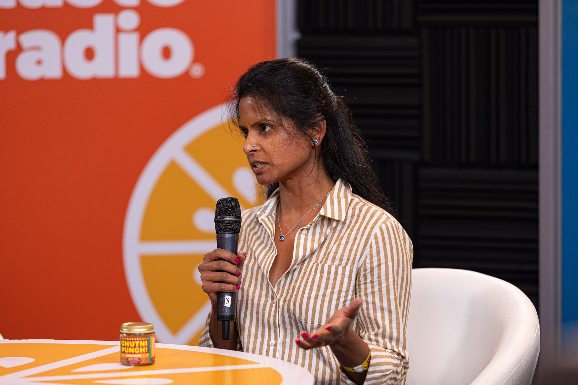 A woman in a striped shirt with a microphone and a small Chutni Punch jar on the orange table.