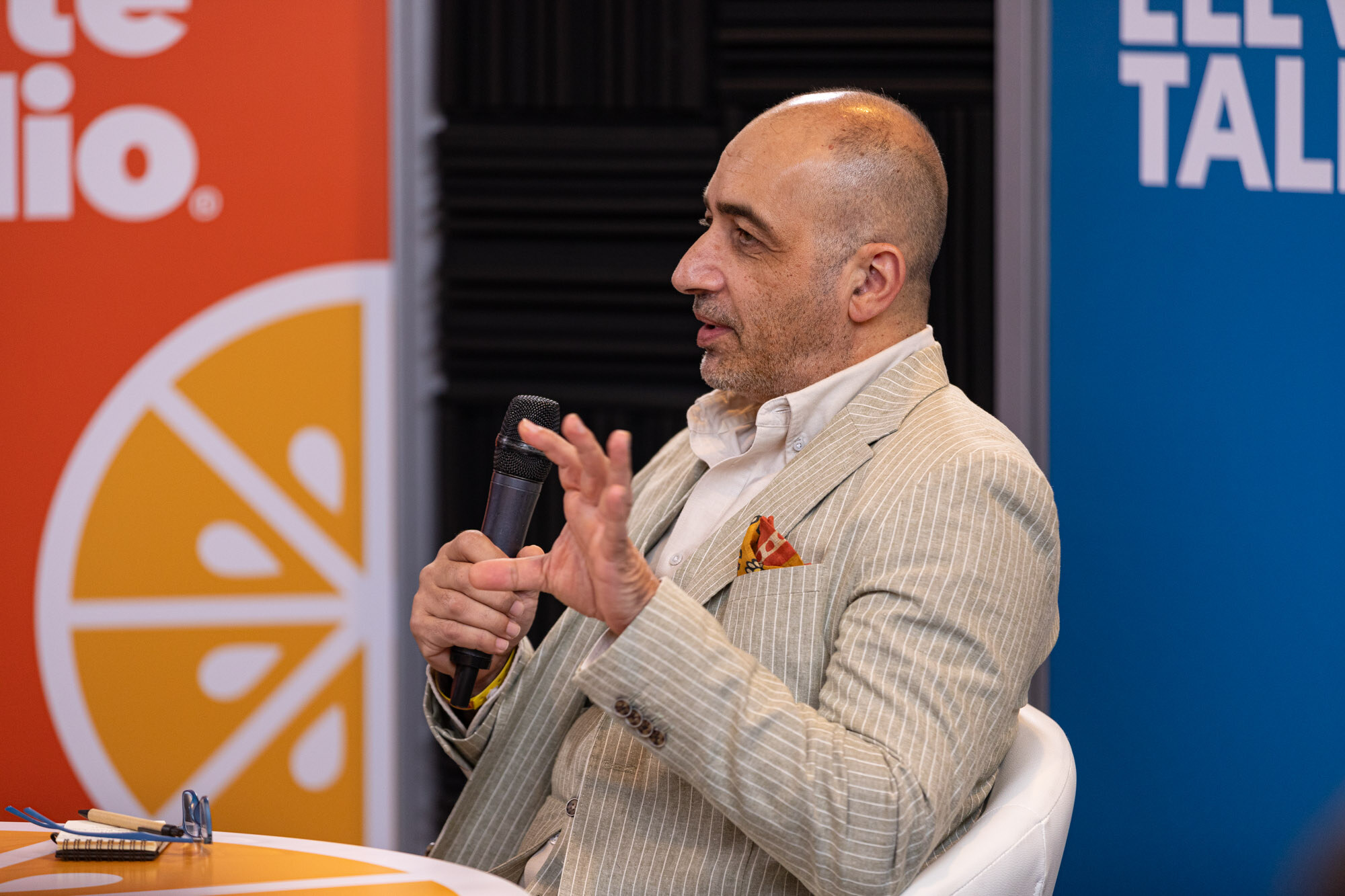 A bald man in a cream pinstripe blazer with a pocket square holding a microphone.