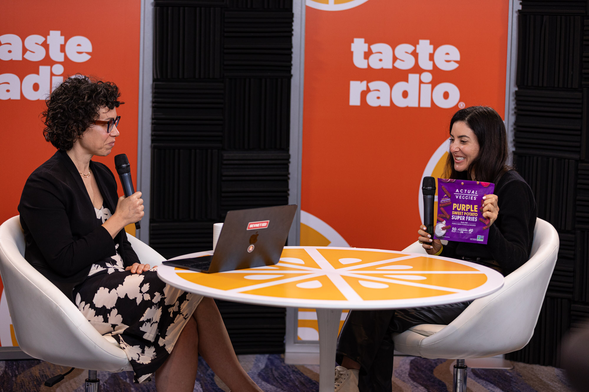 A woman in black holding a purple Actual Veggies bag at the table, with the host across from her.
