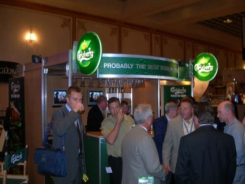 Attendees in suits stand around a booth with green Carlsberg signs. One man in a gray suit drinks a beer, and a blue bag sits on a chair at left.
