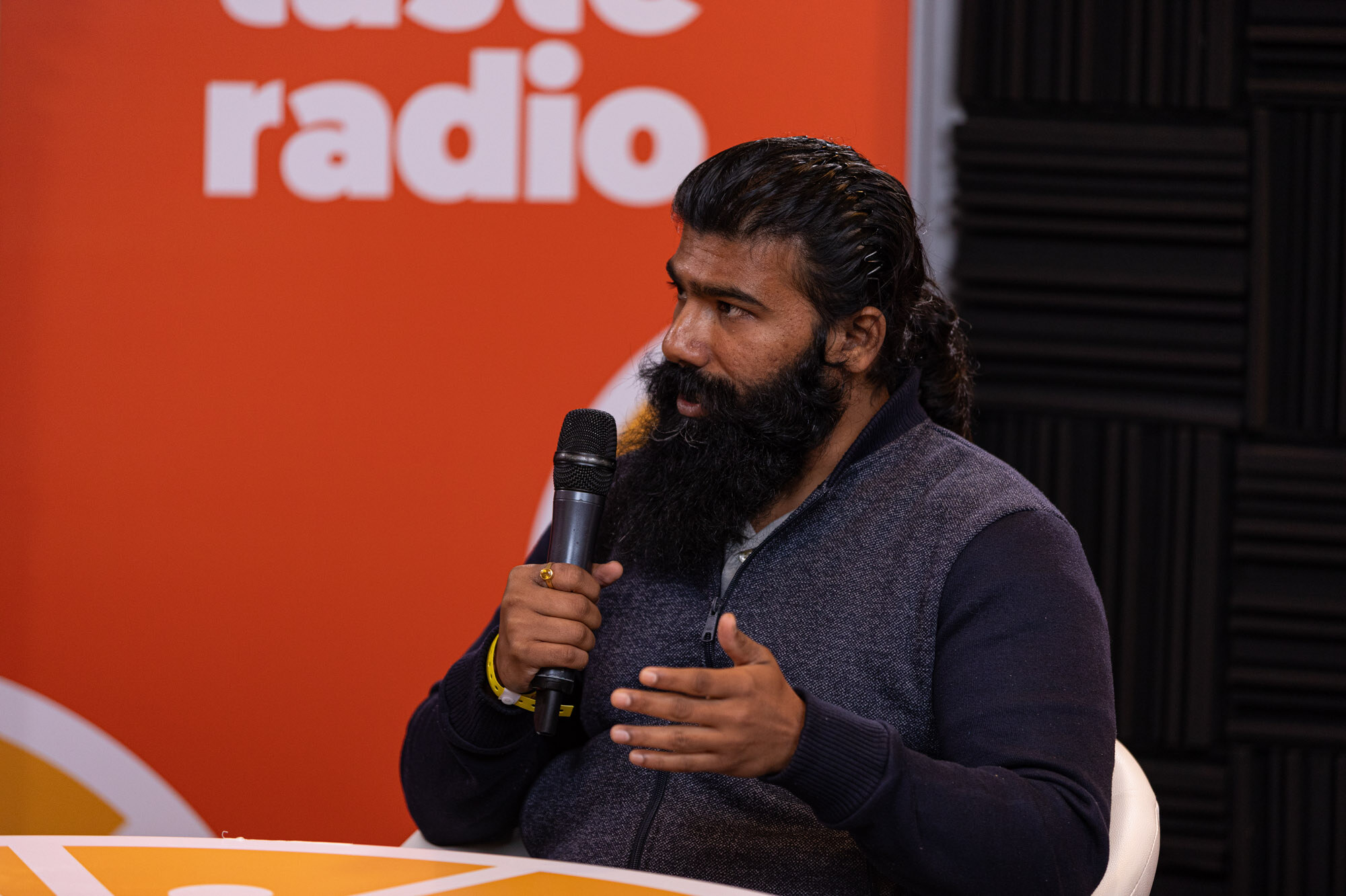 A man with a long beard and ponytail in a navy vest holding a microphone in front of a Taste Radio banner.