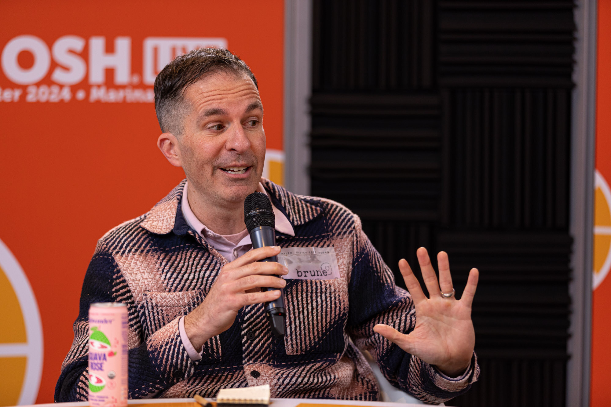 A man in a pink and navy plaid jacket speaking into a microphone beside a canned drink.