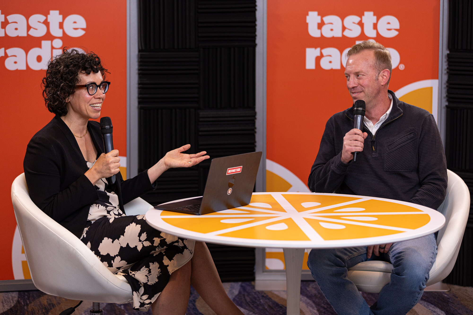 A woman with curly hair in a floral skirt and a man in a navy quarter-zip sitting at a round orange table.