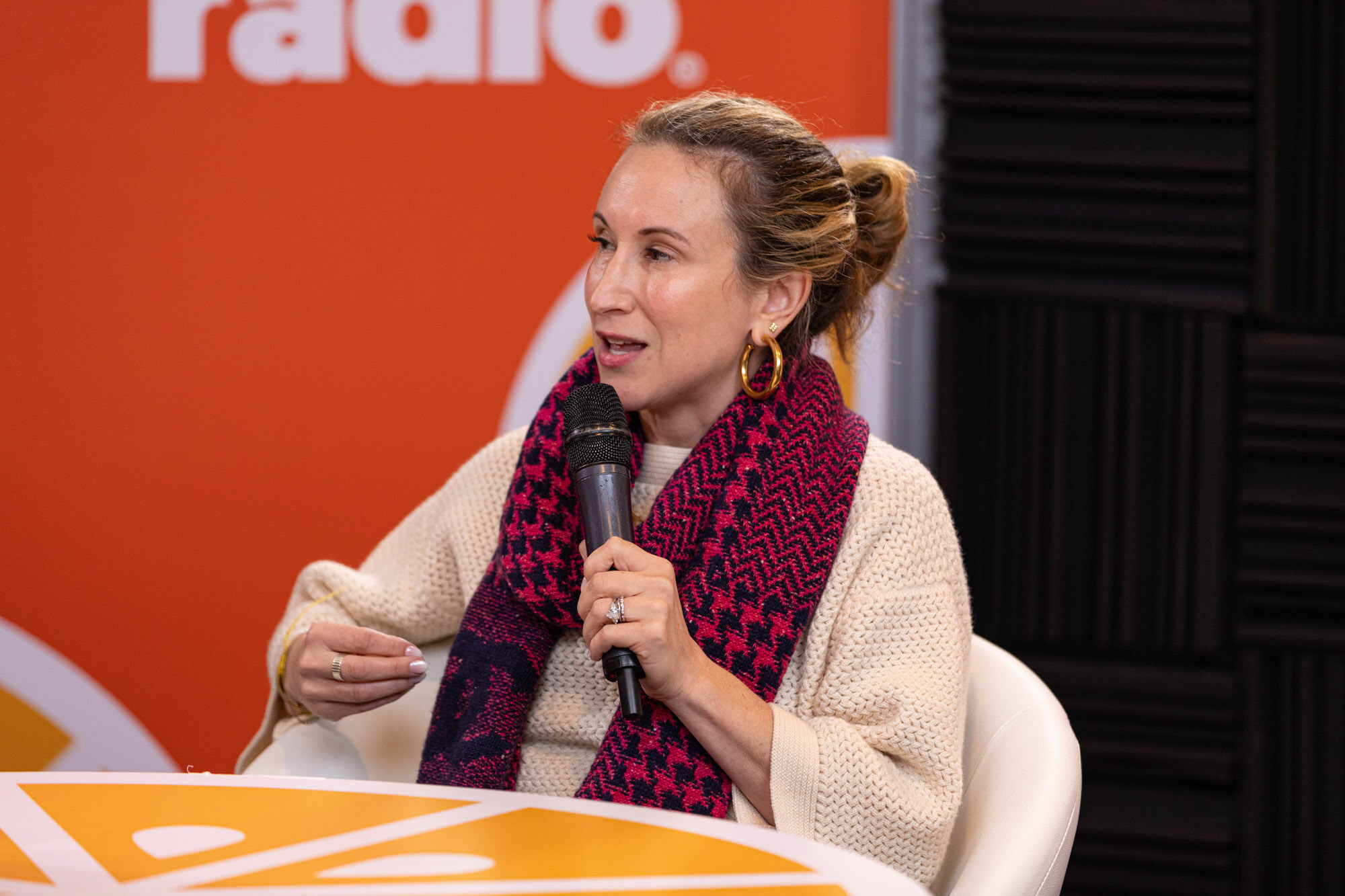 A woman with her hair in a bun, a cream cardigan and a magenta patterned scarf speaking into a microphone.