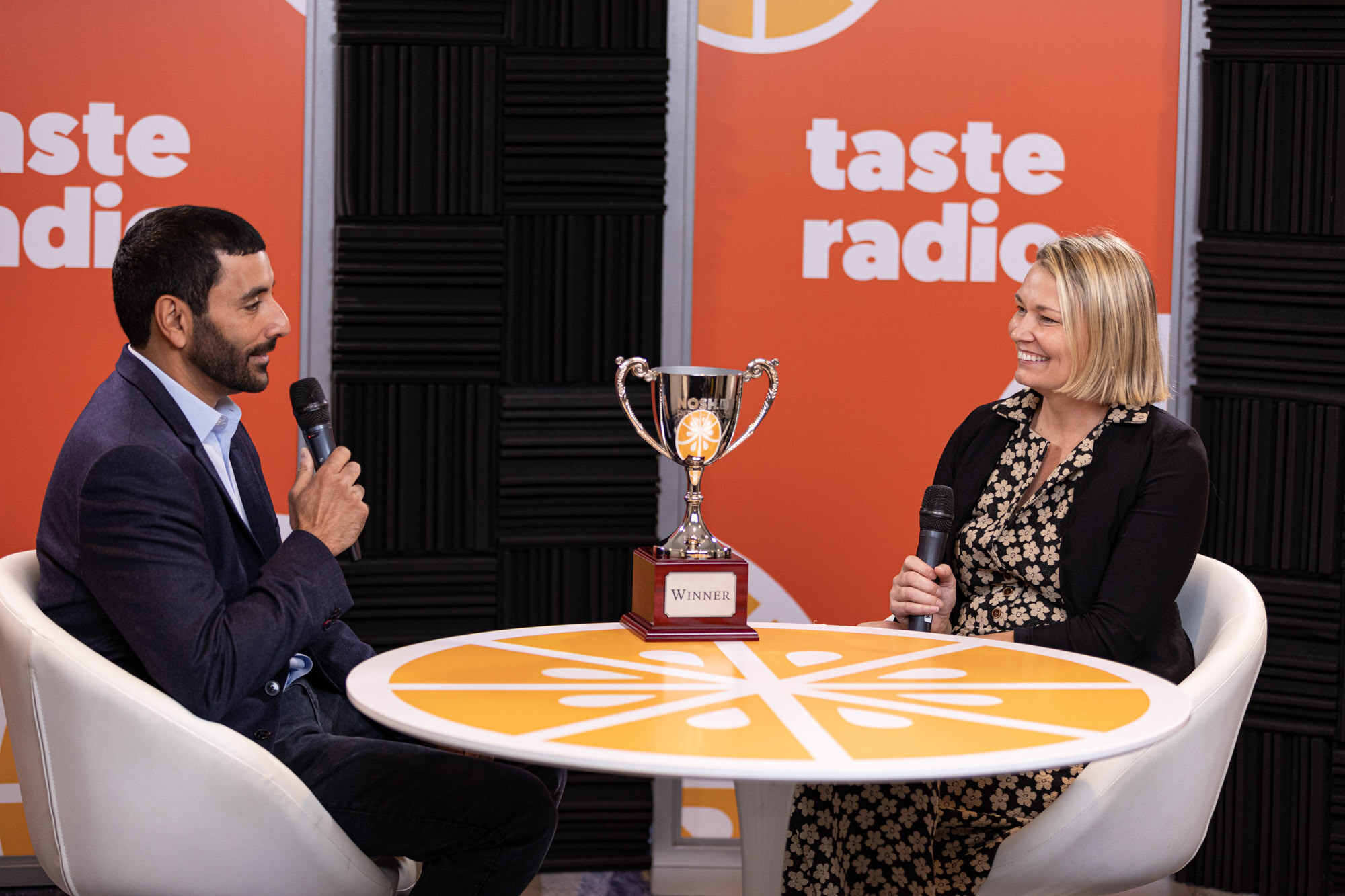 A man in a navy blazer interviewing a woman with a blond bob at a round orange table with a silver Winner trophy.