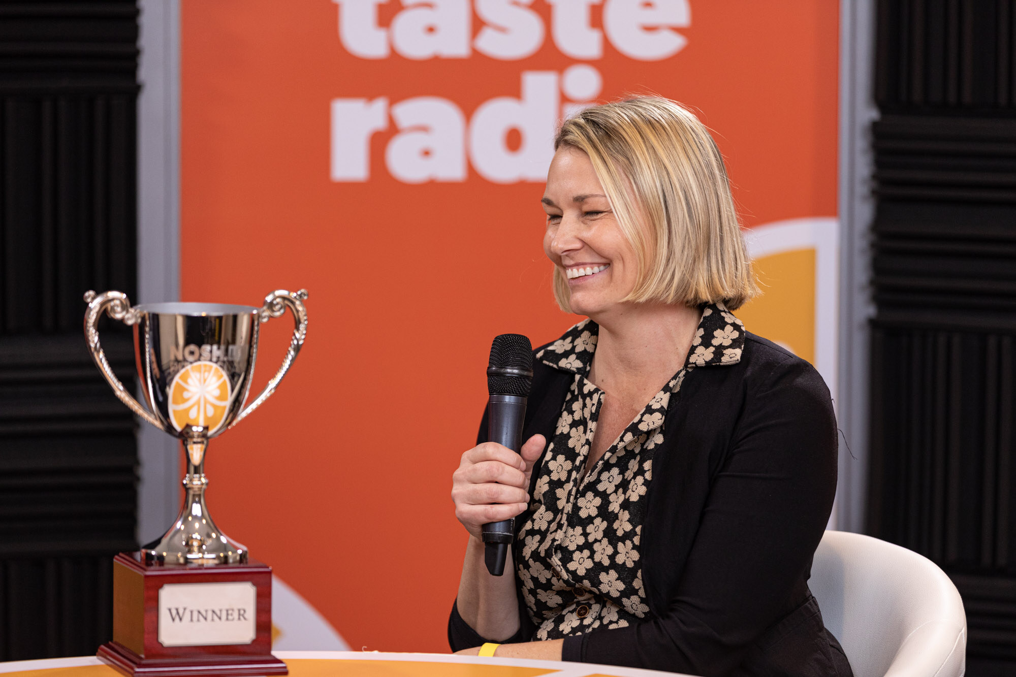A woman with a blond bob in a floral blouse laughing with a microphone next to a silver Winner trophy.