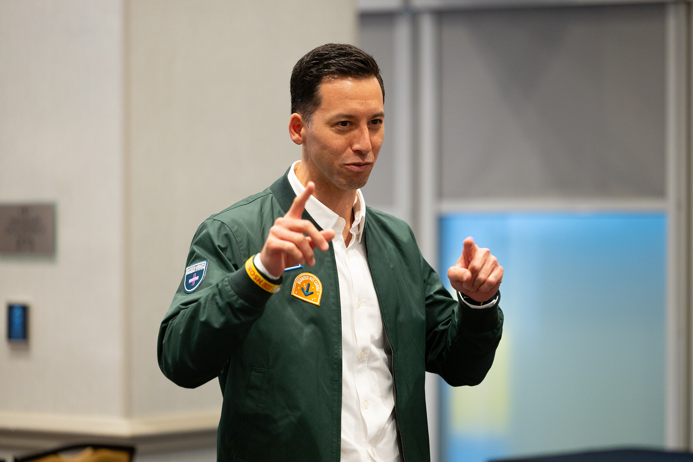 A man in a green bomber jacket and white shirt pointing with both hands.