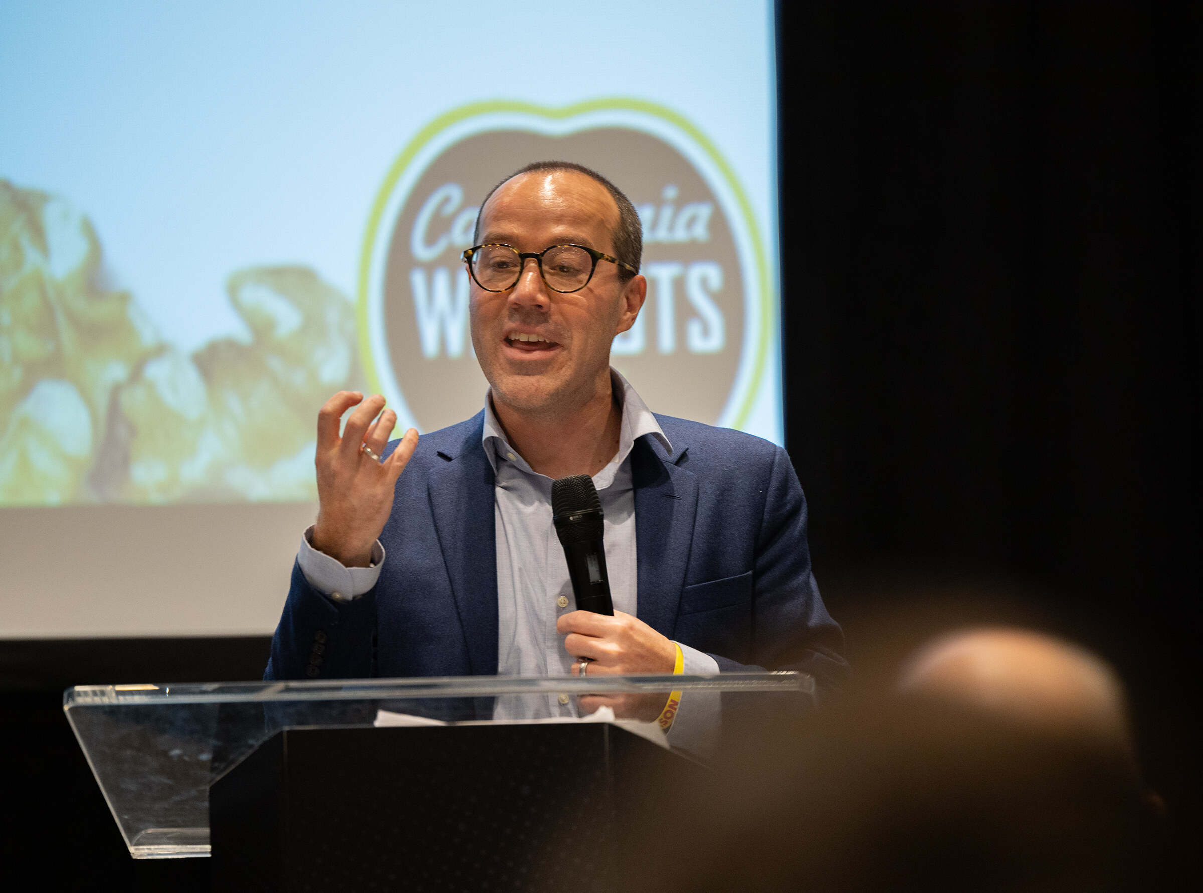 A man in a blue blazer and round glasses speaking with a microphone beside a screen showing California Walnuts.