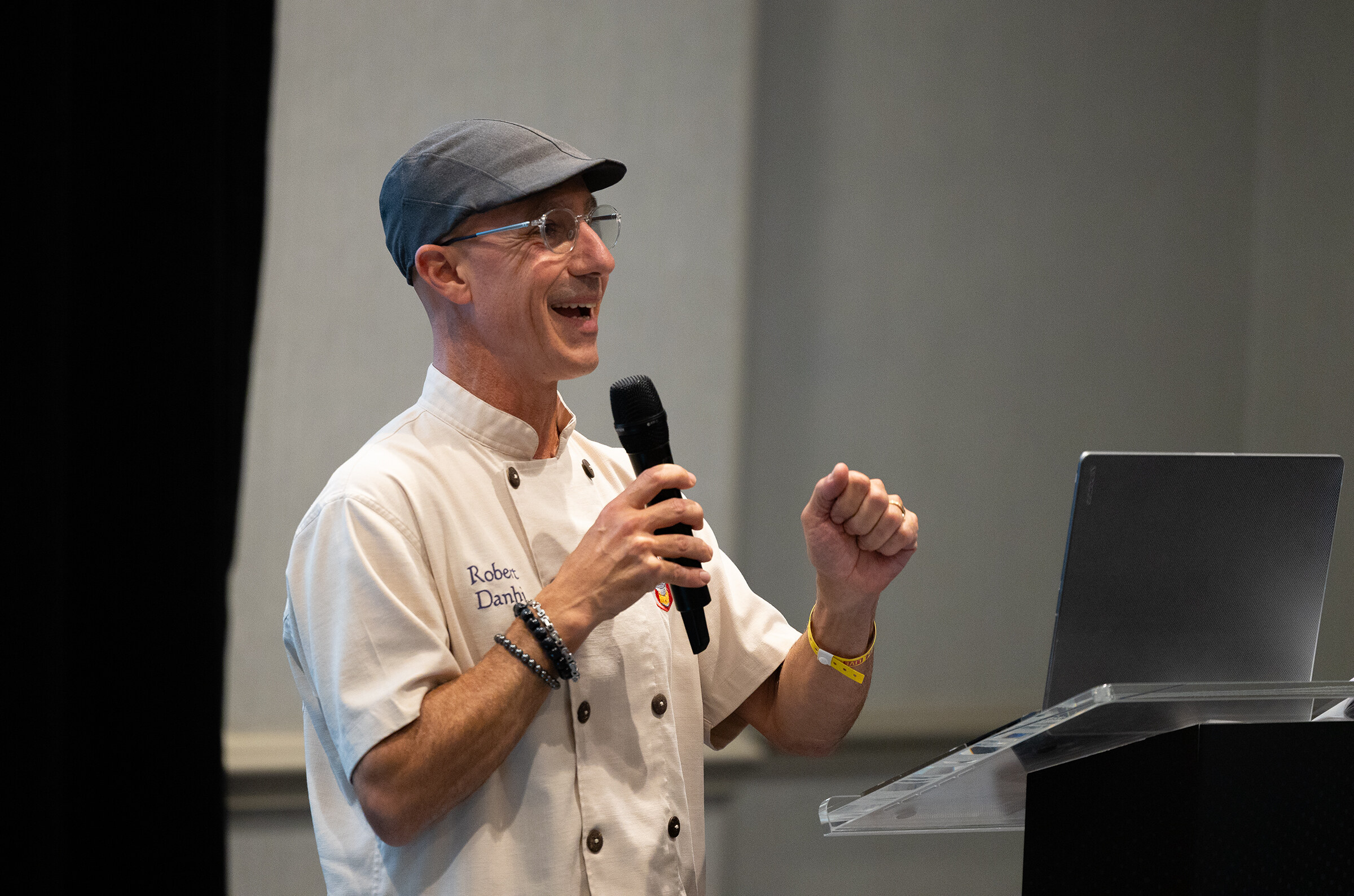 A man in a gray cap and a white chef coat speaking into a microphone at a podium.