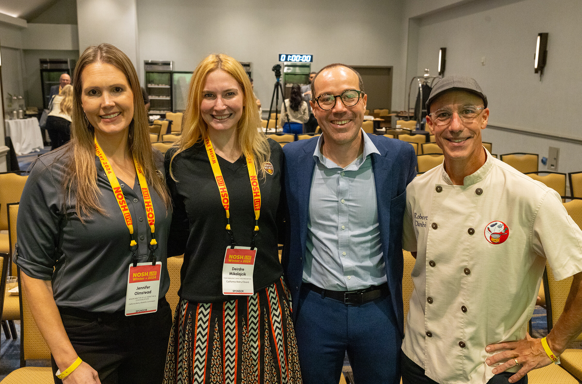 Four people with lanyards standing together smiling in front of rows of chairs.