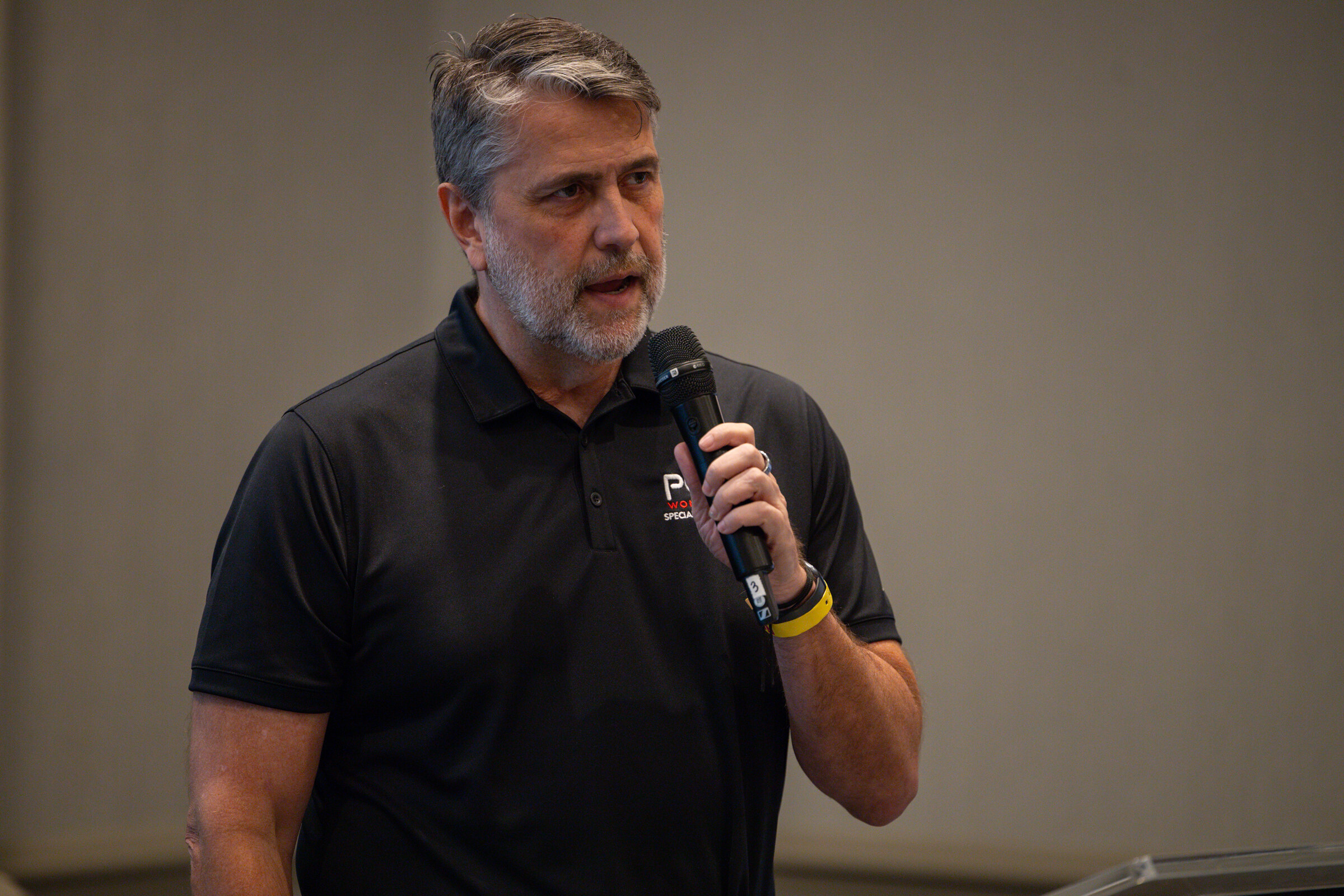 A gray-bearded man in a black POM polo holding a microphone in a meeting room.