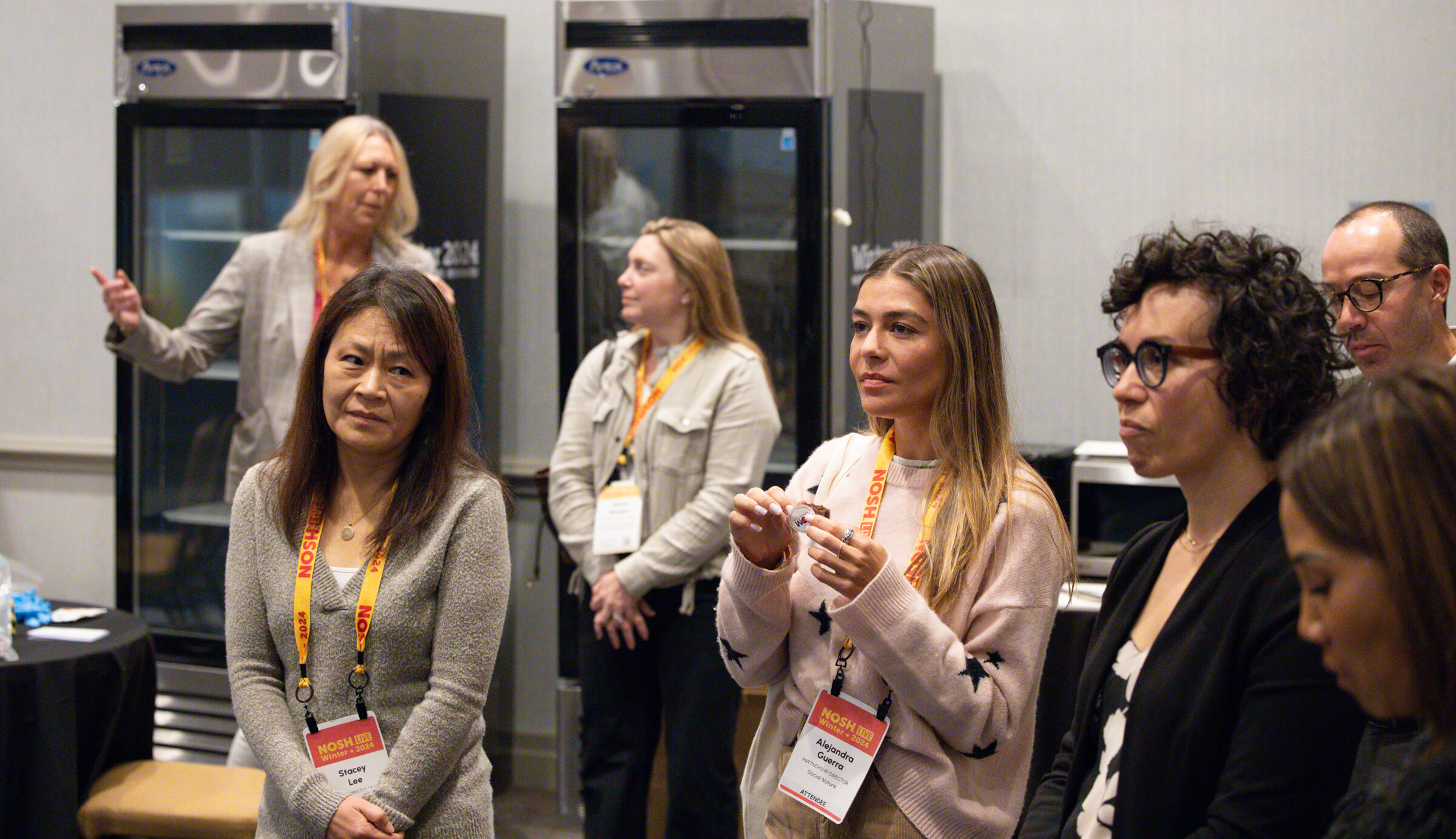 A group of attendees with lanyards standing near glass-door refrigerators listening to a presenter.
