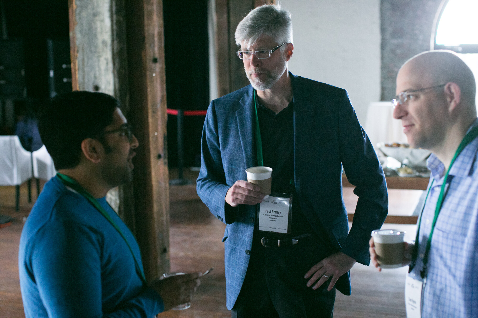 Three men with coffee cups and green lanyards talking beside a wooden post.