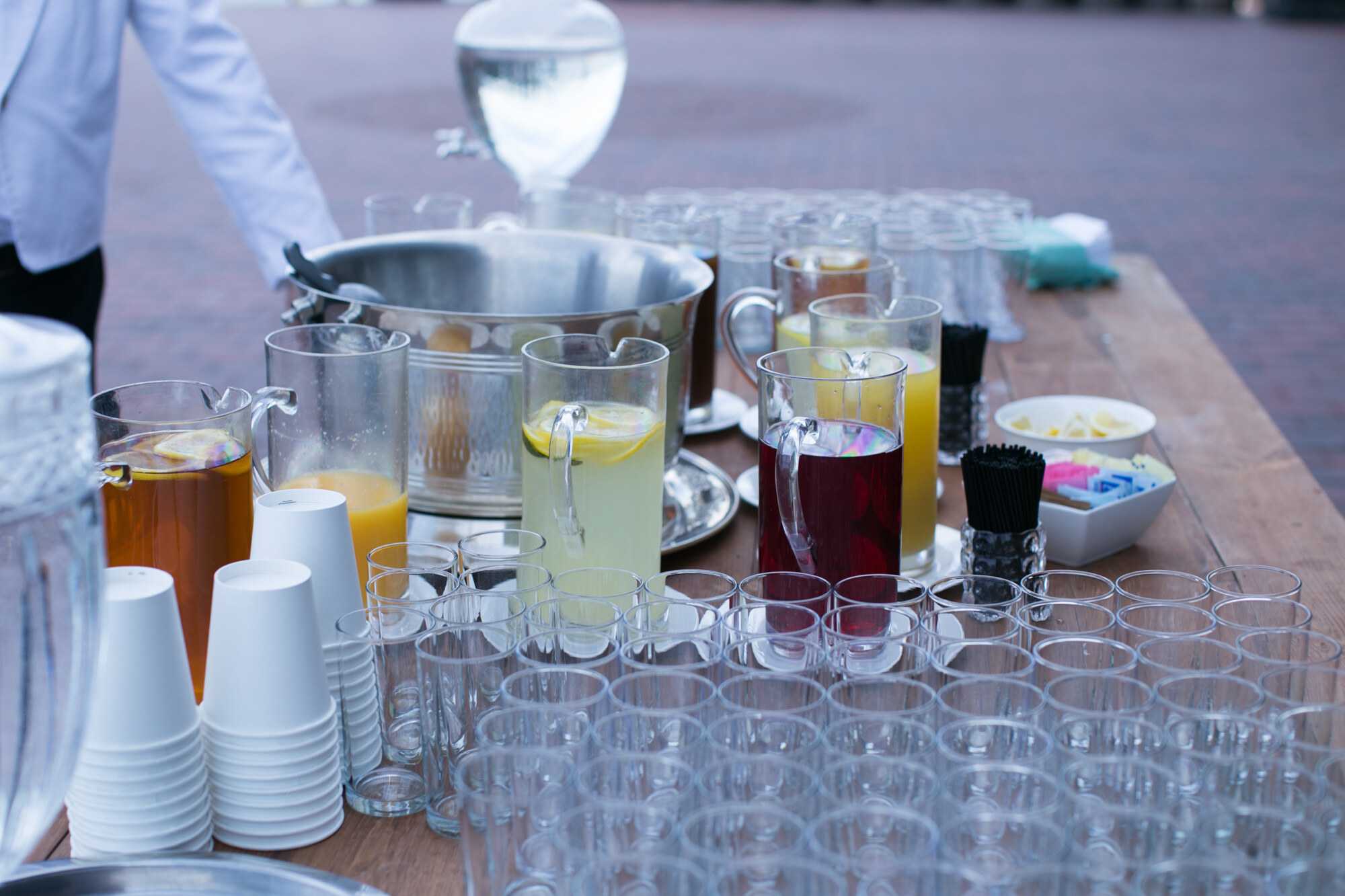 Wooden table with pitchers of lemonade, iced tea and juice and rows of clear glasses.
