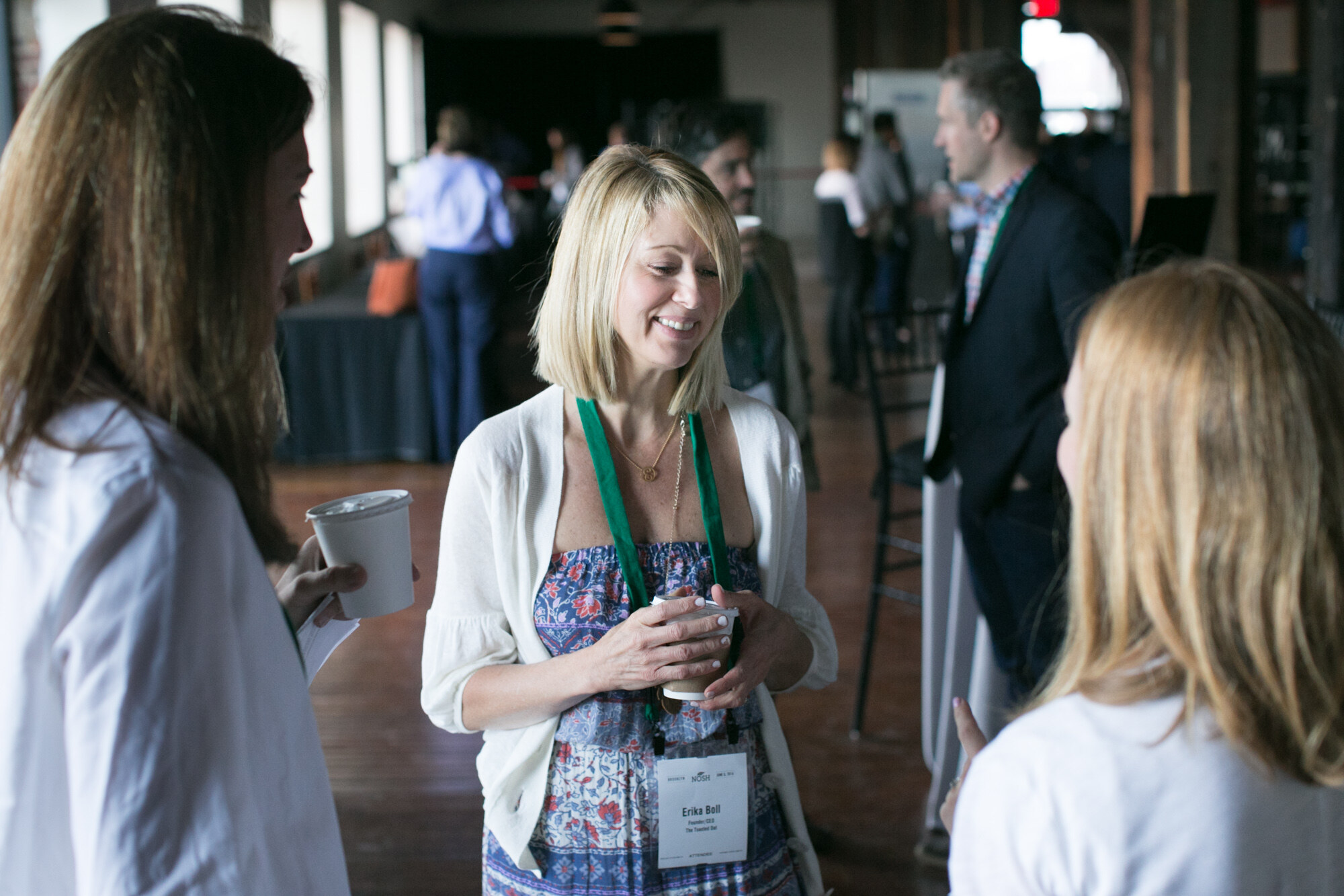 Blonde woman with a coffee cup smiling in conversation with two other attendees in a bright hall.
