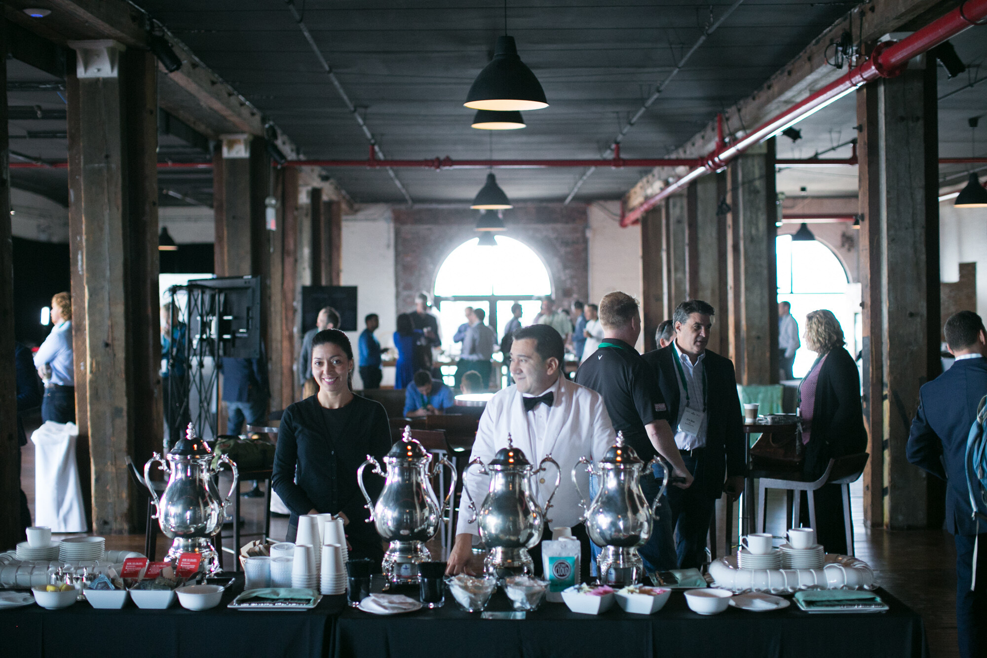 Servers behind a black-draped table with silver coffee urns, cups and trays with attendees in the hall behind.