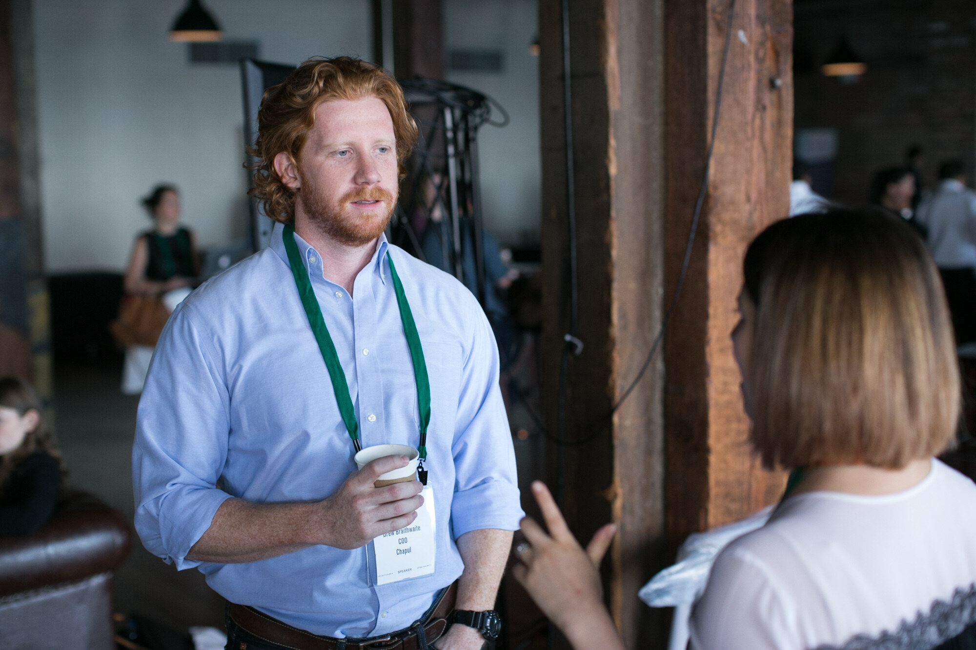 Red-haired man in a light blue shirt and green lanyard holding a coffee cup while a woman in the foreground gestures.
