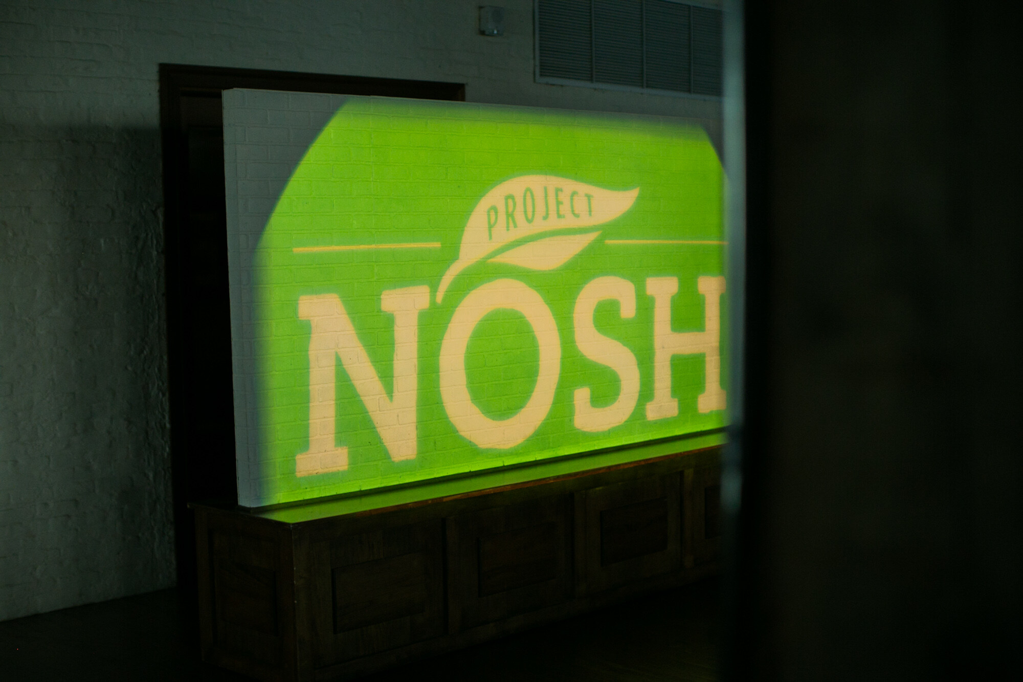 Green projection of the Project NOSH logo on a white brick wall.