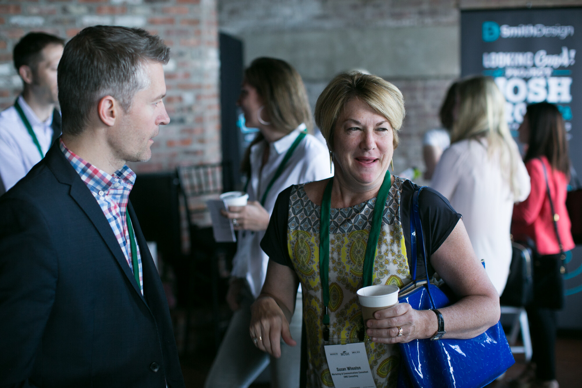 Man in a dark blazer talking with a woman holding a coffee cup, with a Looking Good at Project NOSH banner behind them.