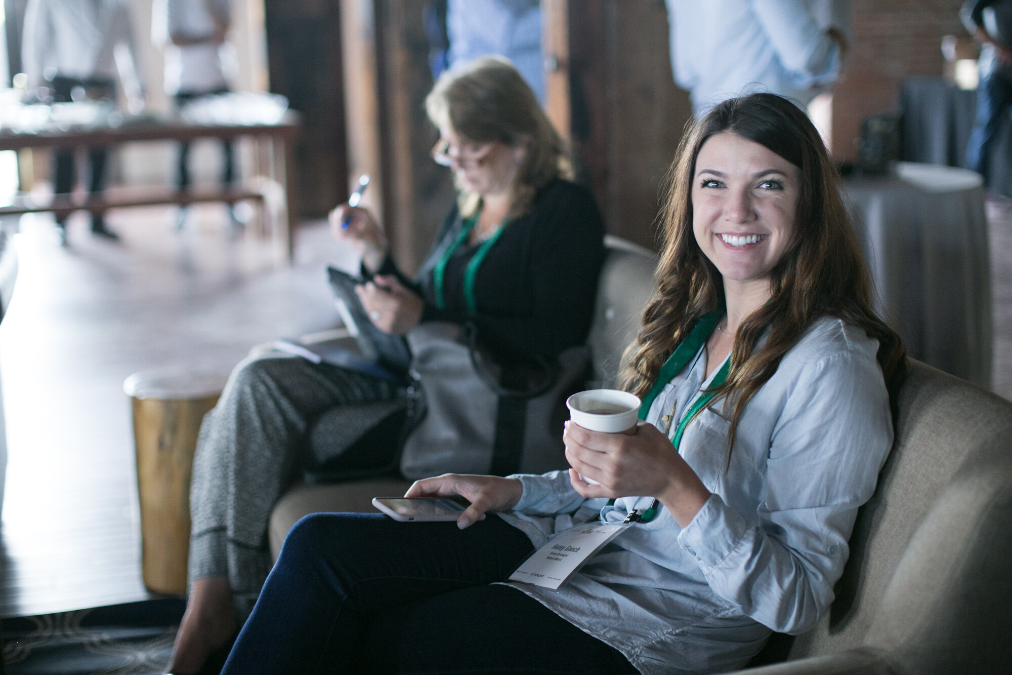 Woman with long brown hair smiling and holding a coffee cup on a sofa, with another woman writing on a tablet beside her.