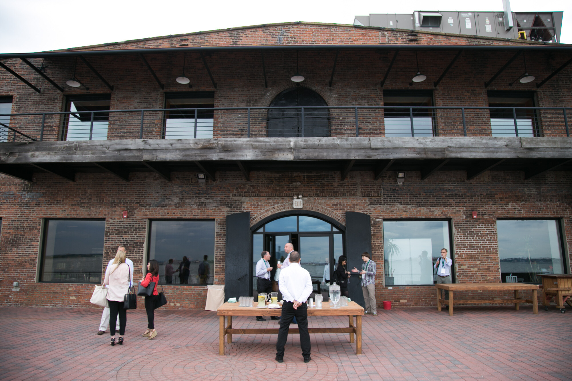 Brick-paved terrace in front of a two-story brick building with a wooden table of drinks and attendees standing outside.