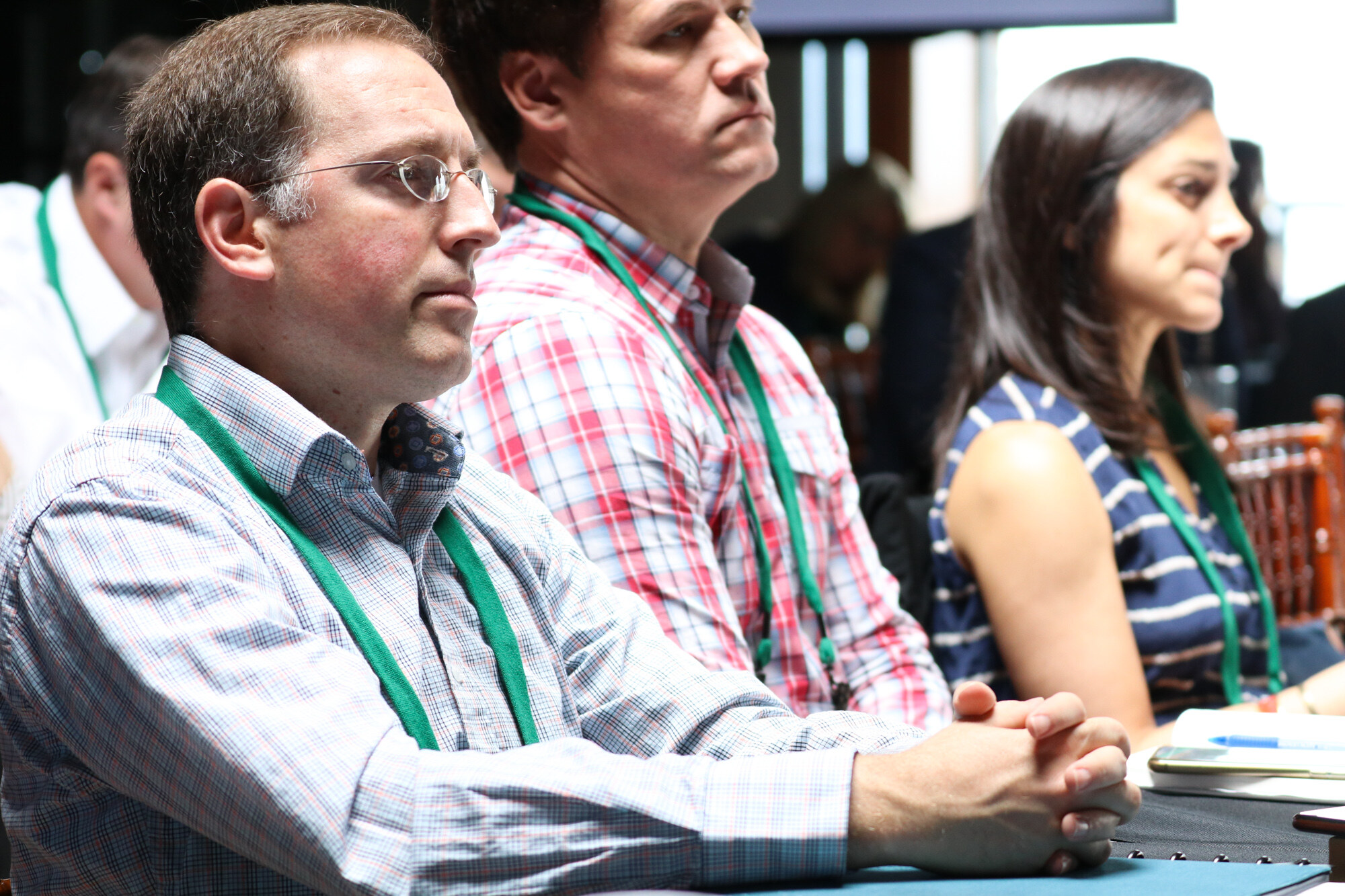 Three attendees in plaid and striped shirts seated at a table, looking toward the stage.