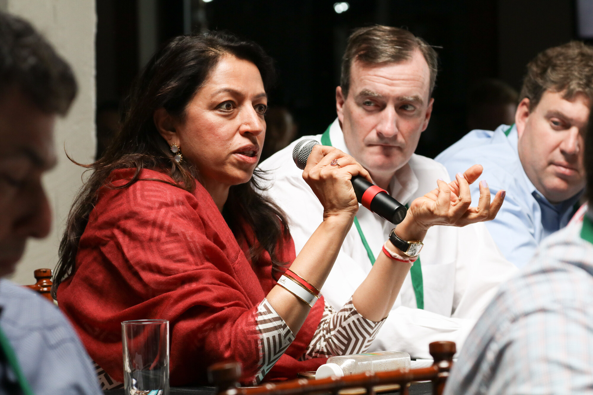 Woman in a red shawl speaking into a microphone at a table, with two men in the background looking on.