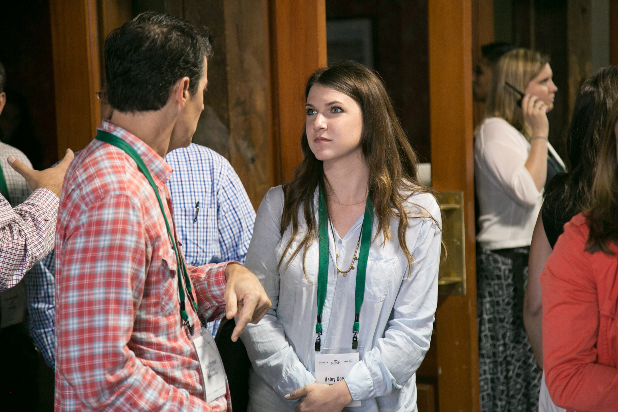 Man in a red plaid shirt talking with a woman with long brown hair beside wood-paneled doors.