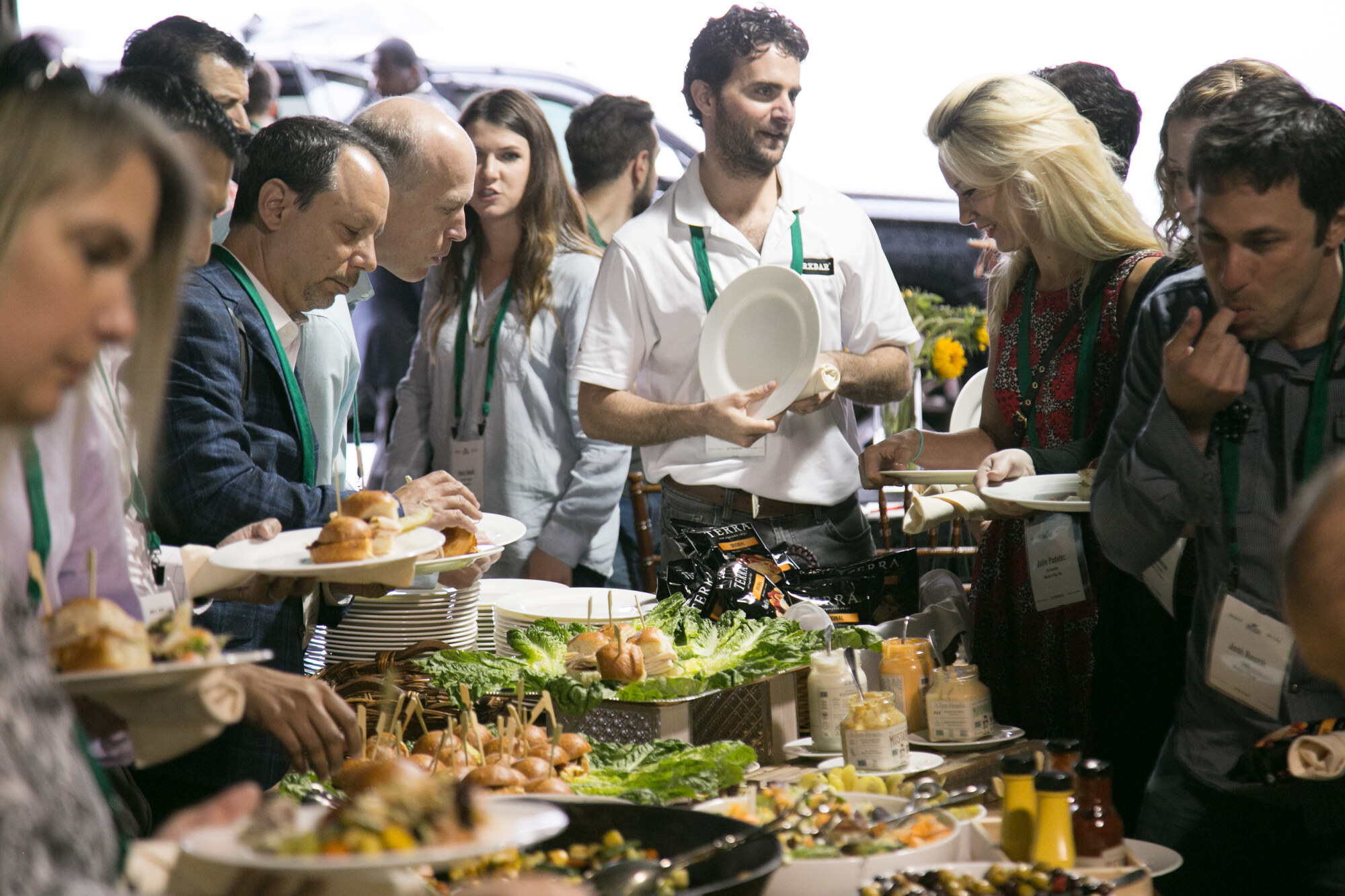 Crowd of attendees with plates at a buffet table with lettuce, sliders, olives and condiments.