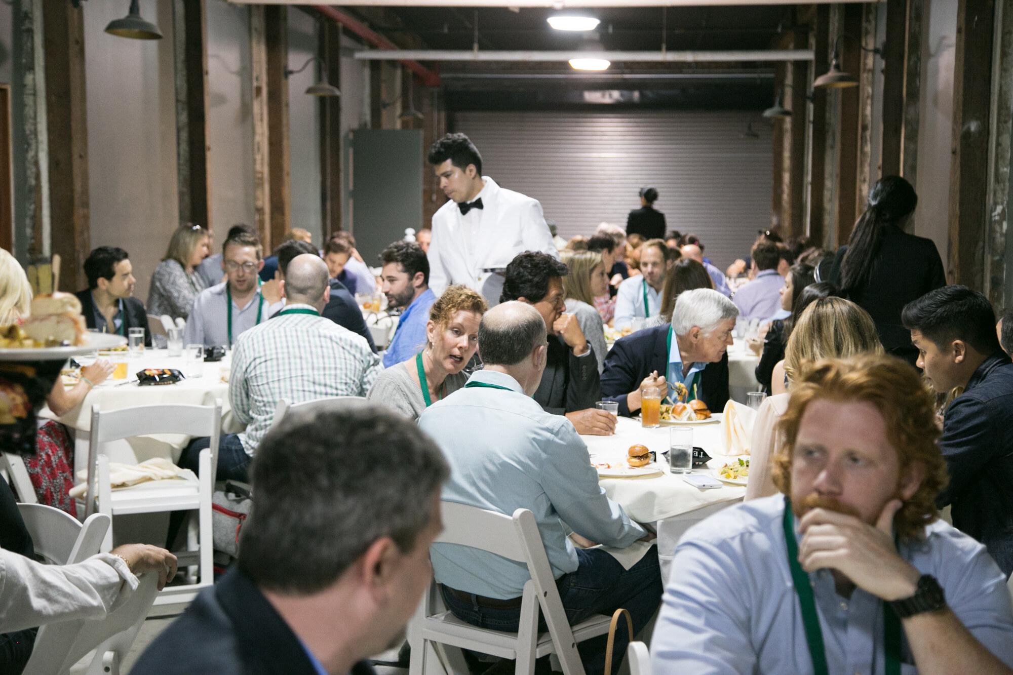 Crowded lunch tables with attendees eating sliders and talking, a waiter in a white jacket standing behind them.