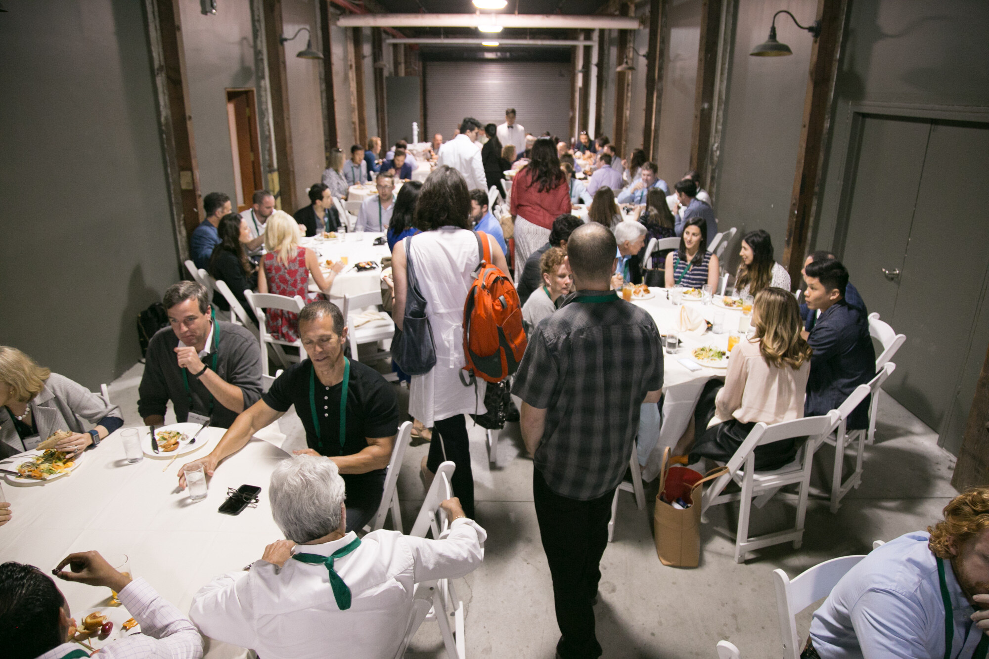 Attendees at round white-clothed tables eating lunch in a long stone-floored hall.