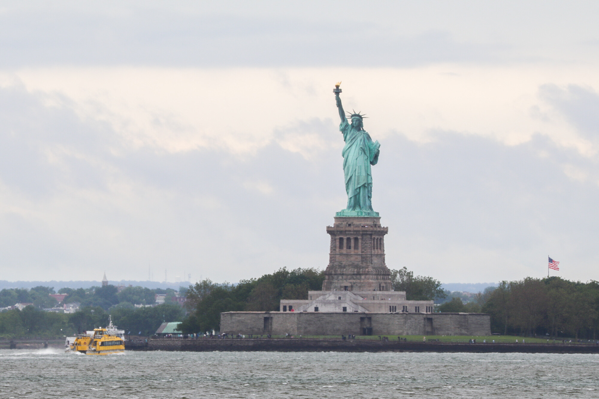 Statue of Liberty on its island under a cloudy sky with a yellow water taxi in the foreground.